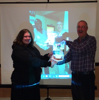 Jim Matthews presents a banner from the Rotary Club of Brusubi to President Gemma Kiddy in front of a photo of Jim receiving it in The Gambia